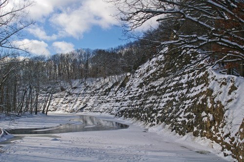 A snowy landscape with a winding river, steep cliffs, and bare trees under a partly cloudy sky.