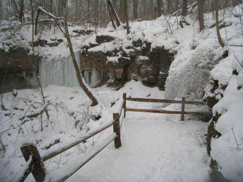 A snowy landscape featuring a frozen waterfall and a wooden fence, surrounded by trees in a winter setting.