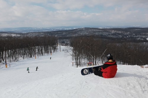 A snowboarder sits on a snowy slope, overlooking a ski trail with trees and hills in the background.