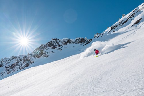 A skier carves through fresh snow on a sunny mountain slope, with bright sunlight shining in the background.