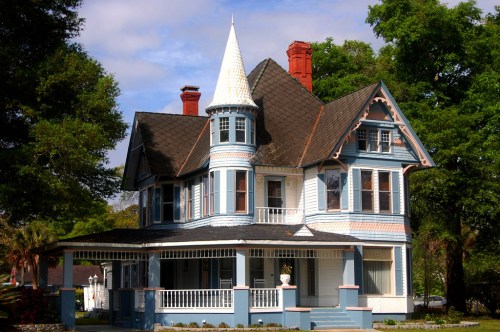 A charming Victorian-style house with a pointed turret, blue siding, and intricate architectural details surrounded by greenery.