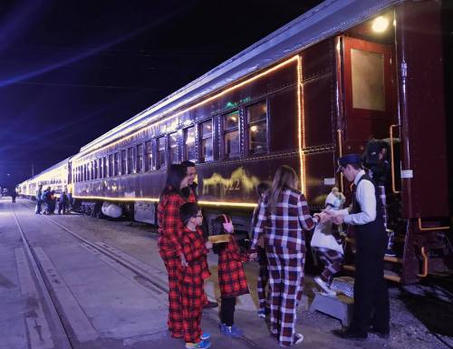 Families in matching pajamas board a festively lit train at night, with a conductor assisting them.