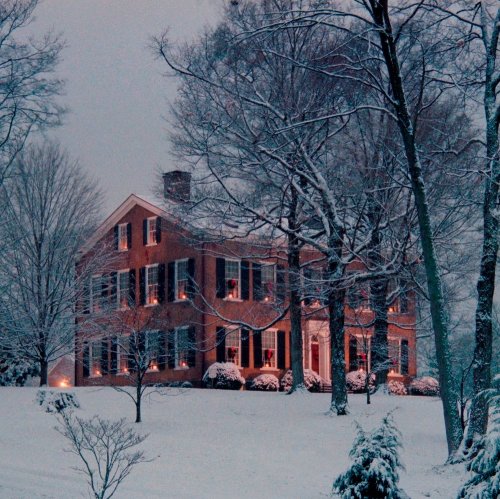 A charming brick house surrounded by snow-covered trees, with warm light glowing from the windows on a winter evening.