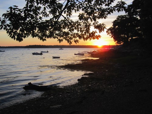 Sunset over a calm lake, with boats anchored and silhouettes of trees framing the scene.