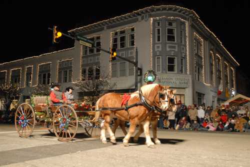 A horse-drawn carriage decorated for the holidays, with riders, passes by a festive building and a crowd watching.