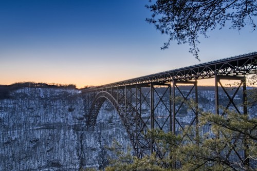 A steel bridge spans a snowy valley at sunset, with trees framing the scene and a clear sky above.