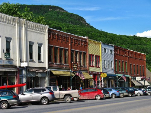 A charming street lined with colorful historic buildings and parked cars, set against a backdrop of green mountains.