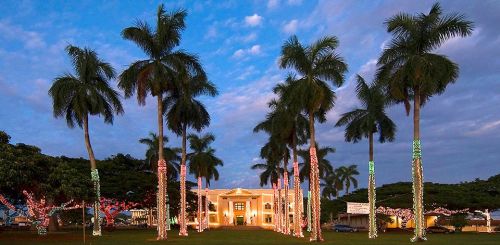 A beautifully lit building surrounded by palm trees, adorned with colorful lights against a cloudy sky.
