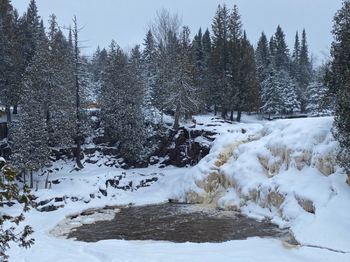A snowy landscape featuring a waterfall cascading over rocks, surrounded by evergreen trees and a wintery scene.
