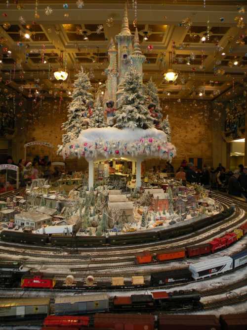 A festive display featuring a miniature train set, snowy trees, and a castle, surrounded by holiday decorations.