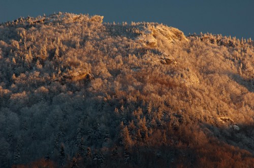 Snow-covered mountain peak illuminated by warm sunlight, surrounded by frosted trees under a clear sky.