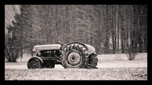 A vintage tractor sits in a snowy field, surrounded by trees in a winter landscape.