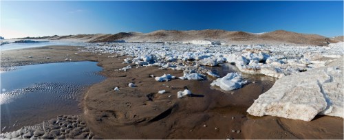 A panoramic view of a sandy beach with ice formations and a clear blue sky in the background.
