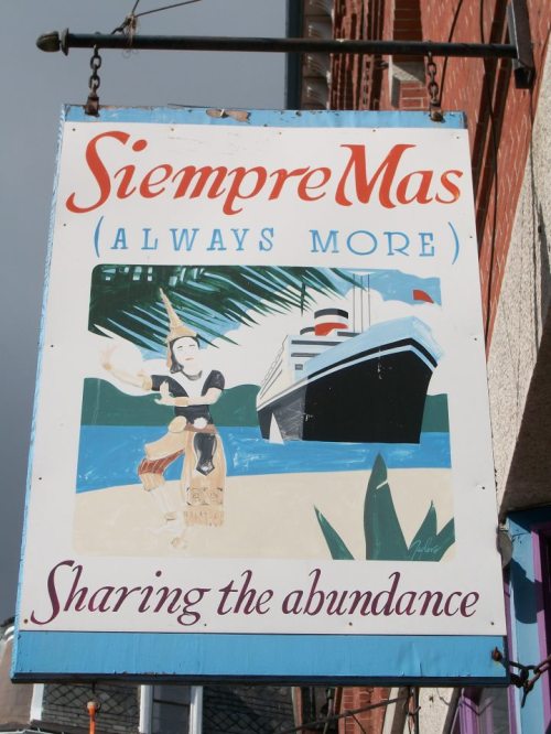 Colorful sign reading "Siempre Más (Always More)" with a beach scene and a ship, promoting abundance.
