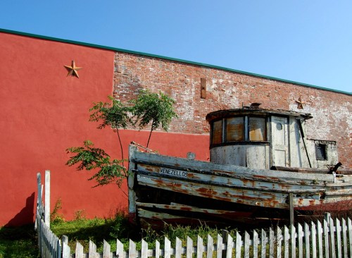 An old, weathered boat named "Venezellos" rests beside a red brick wall with a star, surrounded by greenery and a white fence.