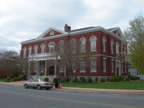 Historic red brick building with white trim, large windows, and a front porch, set on a tree-lined street.