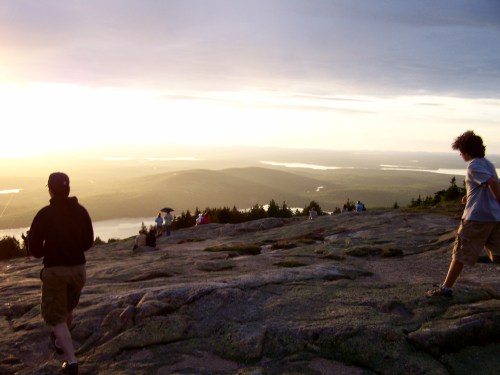 Two people walk on a rocky mountain top at sunset, with others sitting and enjoying the view in the background.