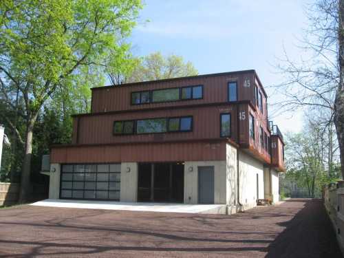 A modern building made from shipping containers, featuring multiple levels and large windows, set in a green environment.