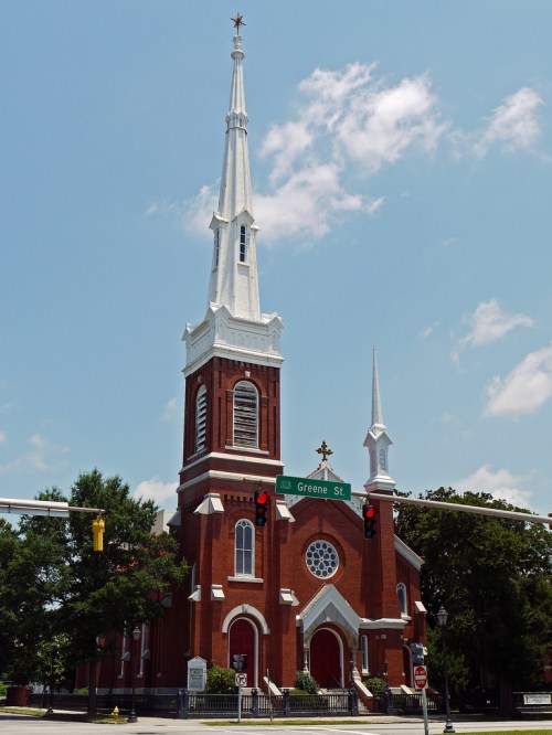 A tall red brick church with a white steeple and cross, set against a blue sky with a street sign nearby.