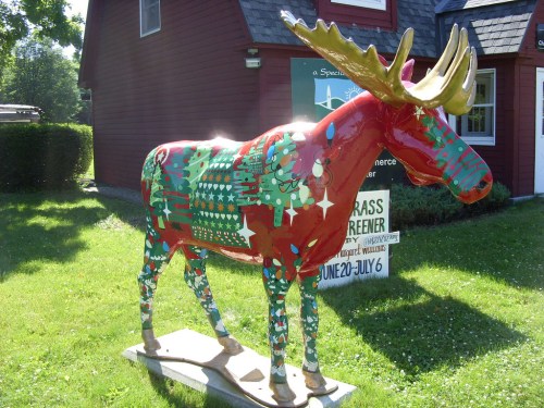 A colorful moose statue decorated with festive patterns stands in front of a red building on a sunny day.