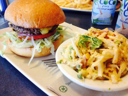 A burger with lettuce, tomato, and cheese next to a bowl of coleslaw on a plate, with drinks in the background.