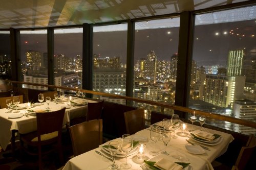 Elegant restaurant setting with tables, glassware, and a stunning city skyline view at night.
