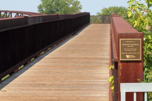 A wooden bridge with a plaque dedicated to Marion Hoffman, surrounded by greenery in a park setting.