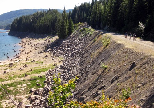 A gravel path along a rocky shoreline, with cyclists riding near a calm lake surrounded by dense trees.