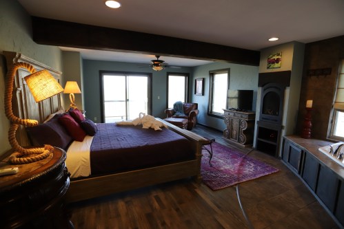 Cozy bedroom with a large bed, armchair, TV, and balcony access, featuring warm lighting and rustic decor.