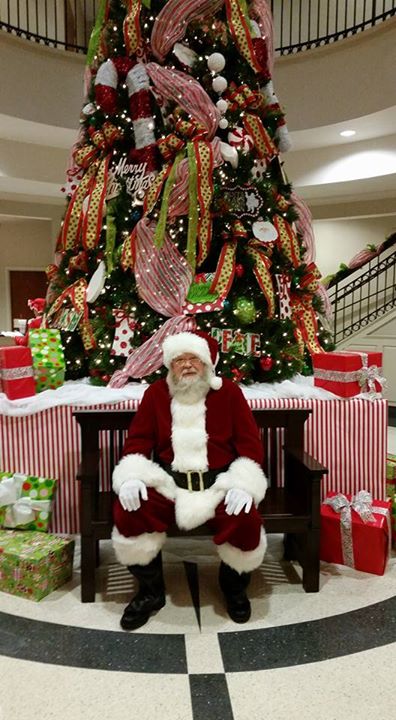 Santa Claus sits on a bench in front of a decorated Christmas tree, surrounded by colorful wrapped gifts.