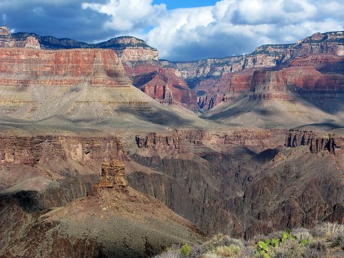 A panoramic view of the Grand Canyon, showcasing layered red rock formations and dramatic cliffs under a cloudy sky.
