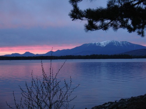 A serene lake at sunset, with mountains in the background and a hint of snow on the peaks, framed by tree branches.