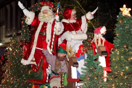 Santa Claus and Mrs. Claus wave from a festive float, surrounded by Christmas trees and holiday lights.