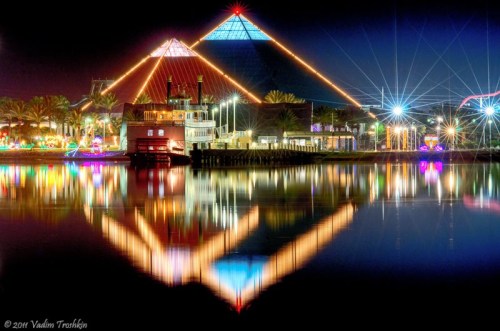 Colorful night scene featuring illuminated pyramids and a boat, reflecting on calm water.