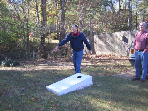 A man throws a bean bag towards a cornhole board in a backyard, while another man watches nearby.