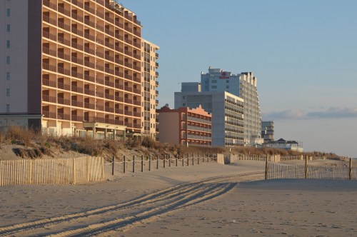 Beach scene with sandy shore, wooden fences, and several tall buildings in the background under a clear sky.