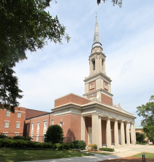 A brick church with a tall steeple, surrounded by trees and a grassy area under a partly cloudy sky.