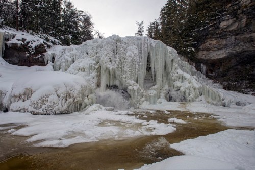 A frozen waterfall surrounded by snow-covered rocks and trees, with icy formations and a partially thawed river below.