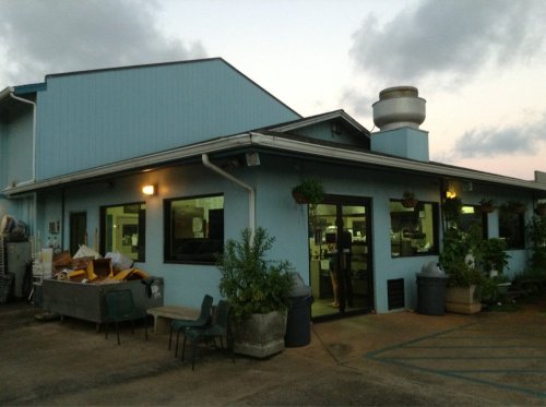A blue building with large windows, outdoor plants, and scattered items outside, under a cloudy sky at dusk.