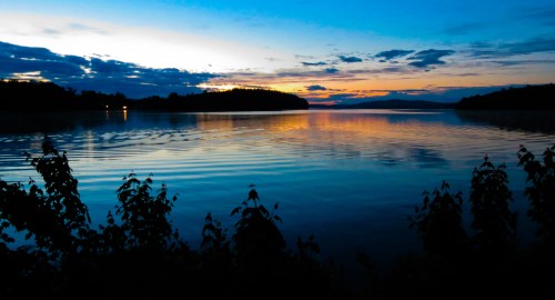 A serene lake at sunset, reflecting vibrant colors in the water, with silhouettes of plants in the foreground.
