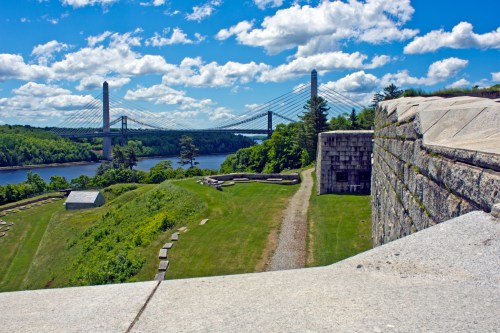A scenic view of a river with a bridge, surrounded by green hills and a historic stone fortification under a blue sky.