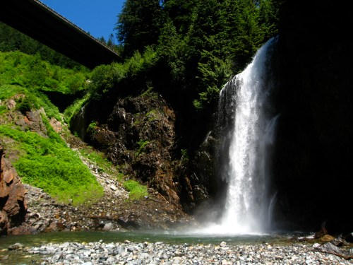 A waterfall cascades down rocky cliffs, surrounded by lush greenery and a bridge overhead under a clear blue sky.