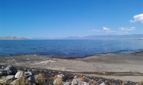 A serene lake scene with calm water, rocky shore, and distant mountains under a clear blue sky.