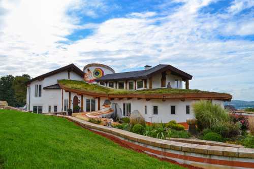 A unique, eco-friendly house with a green roof, colorful design, and landscaped gardens under a cloudy sky.