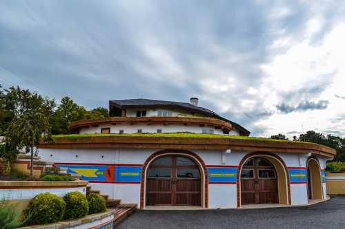 A colorful, round building with a green roof, surrounded by landscaped gardens under a cloudy sky.