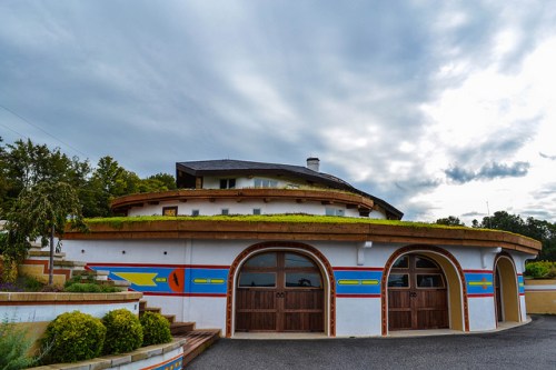 A colorful, round building with a green roof, surrounded by landscaped gardens under a cloudy sky.