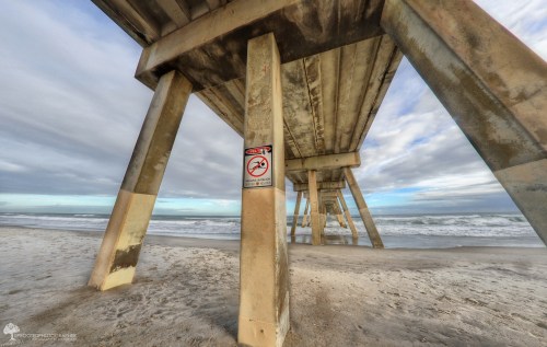 View from under a pier, showing concrete pillars and a no fishing sign, with waves and cloudy sky in the background.