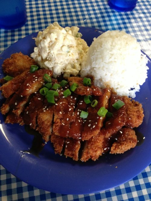 A plate of crispy chicken katsu drizzled with sauce, served with rice and macaroni salad on a blue checkered tablecloth.
