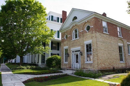A historic brick building next to a large white house, surrounded by green trees and a well-maintained garden.