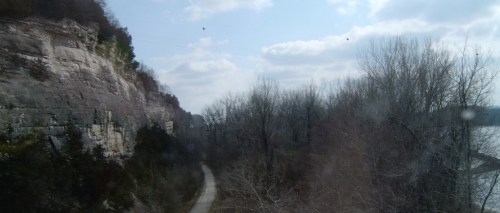 A winding path through a wooded area, flanked by rocky cliffs under a partly cloudy sky.